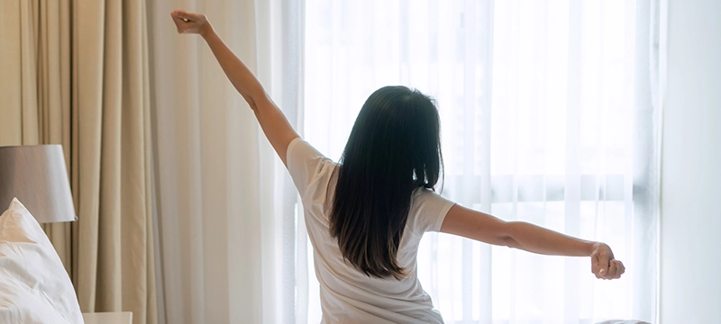 A woman in a white shirt stretches her arms out while awaking in the morning, symbolizing a good night's rest and the importance of good sleep