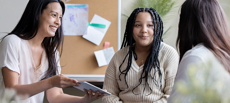 An adolescent student smiles at her mom while her teacher or school psychologist smiles and shows her results on a tablet device, representing how the PDDBI tracks progress over time with RCIs