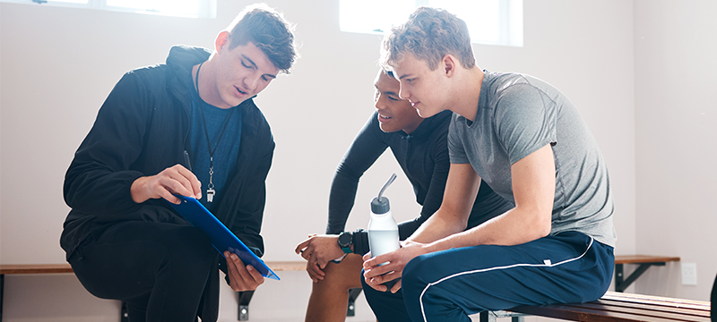 A coach discusses notes with two young players of a sports or athletic team, representative of how the NEO can be used by coaches or sports psychologists to improve team dynamics