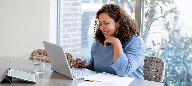 A woman smiles while looking at her laptop in a brightly lit office space with report papers and a tablet device on her desk, representing a school psychologist using AI Report Writer
