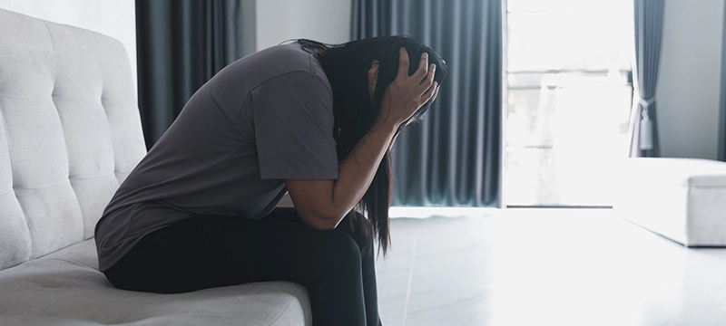 A woman with dark hair sits alone in a brightly lit room clutching her head to represent racing thoughts and Schizophrenia