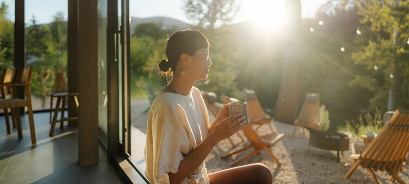 A beautiful woman with her hair pulled back smiles while holding a mug and looking out on a sunny wooded landscape, symbolizing good mental health and relaxation as well as connecting with nature